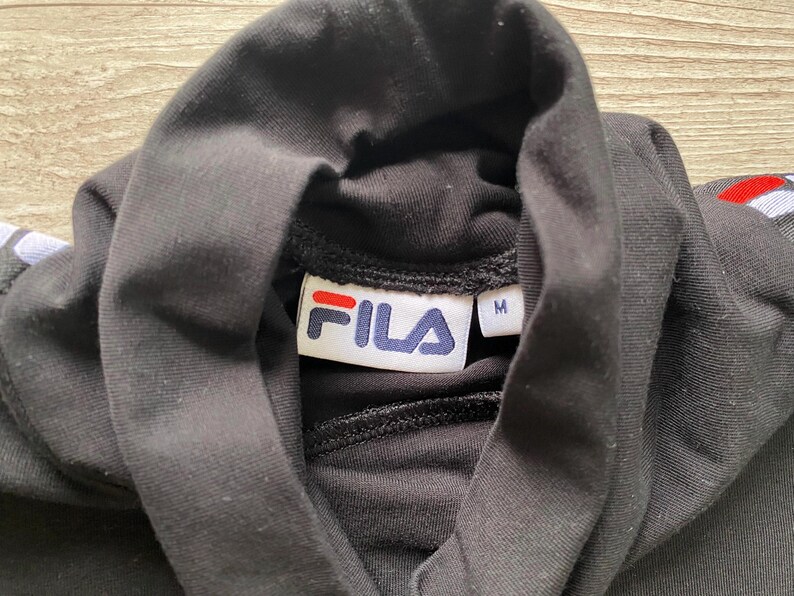 Puede incluir: Una camiseta negra de cuello alto con una etiqueta interior blanca con el logotipo de FILA en rojo y azul y una etiqueta separada que indica la talla M. La prenda presenta detalles de rayas rojas, blancas y azules en las costuras de los hombros.