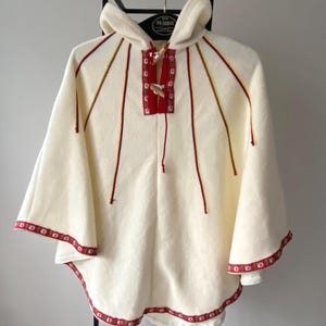 Puede incluir: Poncho con capucha de color crema con detalles bordados en rojo y amarillo. El poncho presenta un ribete rojo con motivos florales en los bordes y un panel rojo con cierres de madera en el cuello. La capucha y las mangas son amplias y fluidas.
