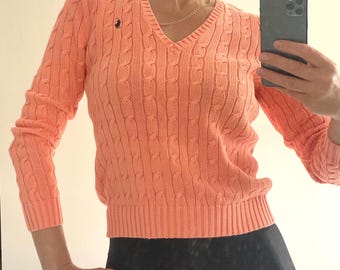 Peut inclure: Un pull à col en V et manches longues de couleur pêche, doté d'un motif en tricot torsadé vertical et d'un petit logo brodé bleu foncé sur la poitrine gauche. Le pull présente des poignets et un ourlet côtelés pour une coupe ajustée.