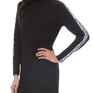 Puede incluir: Un vestido negro de manga larga con cuello alto. Las mangas presentan una franja vertical blanca que va desde el hombro hasta el pu&ntilde;o, decorada con el logotipo de Fila repetido en rojo y azul marino a lo largo de toda la franja.