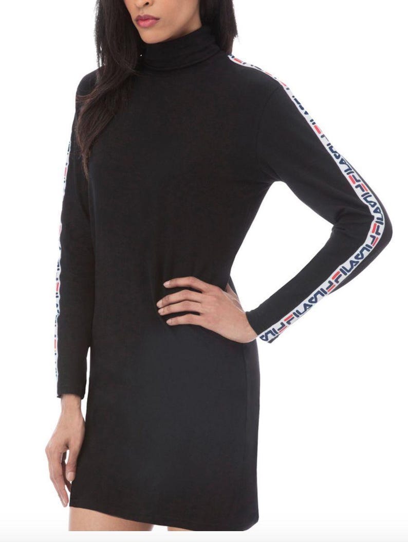 Puede incluir: Un vestido negro de manga larga con cuello alto. Las mangas presentan una franja vertical blanca que va desde el hombro hasta el pu&ntilde;o, decorada con el logotipo de Fila repetido en rojo y azul marino a lo largo de toda la franja.