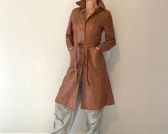 Peut inclure: Un trench-coat en cuir marron clair doté d'un col pointu, d'une fermeture boutonnée sur le devant et d'une ceinture à nouer assortie à la taille. Il est porté avec un pantalon ample gris clair, des bottines noires et des lunettes de soleil.