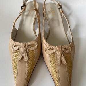 May include: A pair of beige leather slingback heels with pointed toes. The vamp features a gold snakeskin pattern and a bow detail. The heels have a buckle closure and a small heel. The shoes are likely for formal occasions.
