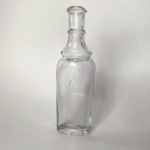 以下が含まれることがあります： 細い首とフレアトップの透明なガラス瓶。ボトルには「1841」の数字と円形のエンブレムがエンボス加工されています。ボトルは空で、白い表面に置かれています。