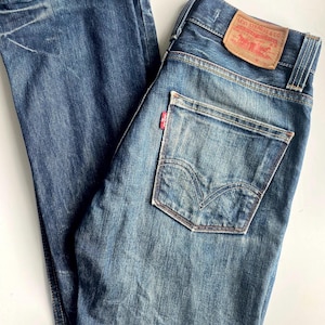 Könnte beinhalten: Blaue Jeans aus Denim mit klassischem Fünf-Taschen-Design. Die Jeans haben eine verwaschene Optik, ein rotes Levi's-Etikett auf der Rückseite und ein kleines rotes Etikett auf der Gesäßtasche. Die Jeans sind gefaltet und flach hingelegt.