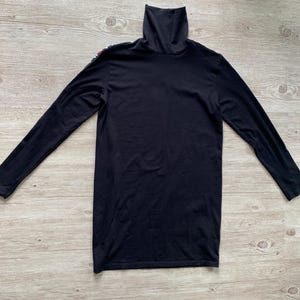 Puede incluir: Una camiseta negra de manga larga con cuello alto y un detalle de raya vertical fina en el hombro derecho. La prenda est&aacute; confeccionada en tela negra lisa e incluye un cuello alto que llega hasta la barbilla, con mangas largas.