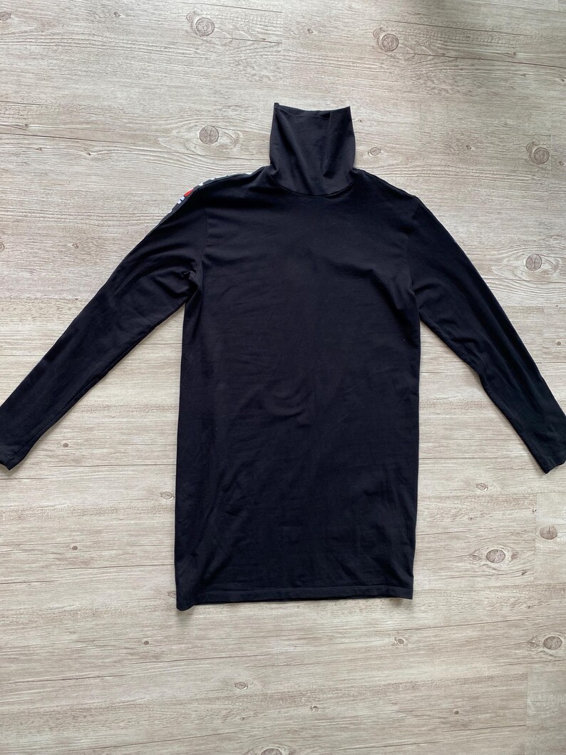Puede incluir: Una camiseta negra de manga larga con cuello alto y un detalle de raya vertical fina en el hombro derecho. La prenda est&aacute; confeccionada en tela negra lisa e incluye un cuello alto que llega hasta la barbilla, con mangas largas.