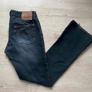 以下が含まれることがあります： 色あせた外観の濃い色のデニムジーンズ。ジーンズは、クラシックな5ポケットのデザイン、赤いLevi'sタグ、背面の革パッチが特徴です。ジーンズは木製の表面に平らに置かれています。