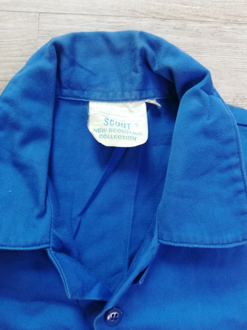Pode incluir: Uma camisa azul de colarinho com uma etiqueta branca que diz "Scout New Scoutline Collection".