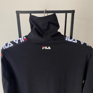 Puede incluir: Una camiseta negra de manga larga con cuello alto y el logotipo de Fila en blanco y rojo en el centro del pecho. Los hombros y las mangas est&aacute;n acentuados con una banda blanca de marca que muestra el nombre Fila en un patr&oacute;n repetitivo.