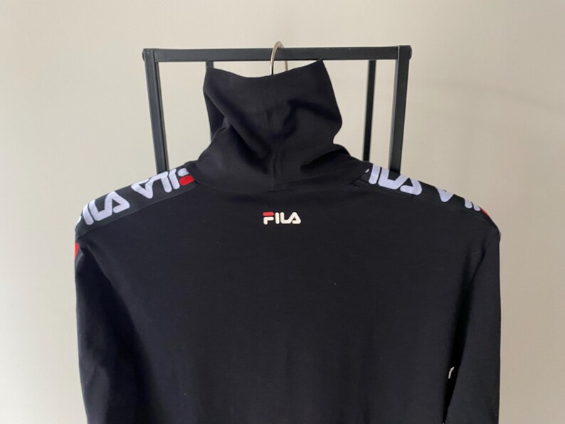 Puede incluir: Una camiseta negra de manga larga con cuello alto y el logotipo de Fila en blanco y rojo en el centro del pecho. Los hombros y las mangas est&aacute;n acentuados con una banda blanca de marca que muestra el nombre Fila en un patr&oacute;n repetitivo.