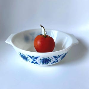 May include: A white milk glass bowl with two side handles, featuring a repeating blue and green floral pattern around the exterior. A single red tomato with a green stem is placed inside the centre of the bowl against a plain white background.