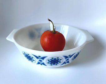 Peut inclure: Un bol en verre opalin blanc avec deux poignées latérales, orné d'un motif floral bleu et vert répétitif sur l'extérieur. Une tomate rouge avec une tige verte est placée au centre du bol sur un fond blanc uni et neutre.