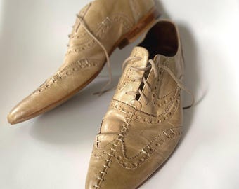 Peut inclure: Une paire de chaussures richelieu en cuir beige avec un bout pointu, une fermeture à lacets et des détails brogues décoratifs. Les chaussures présentent une couture apparente sur le centre du bout et un petit talon à la finition lisse.