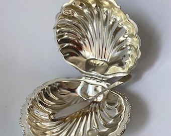 Peut inclure: Un plat en métal argenté en forme de coquillage avec un couvercle à charnière qui s'ouvre pour révéler un intérieur concave avec des détails nervurés. Un petit couteau à tartiner en métal argenté assorti repose dans la partie inférieure.