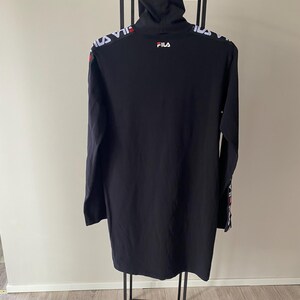 Puede incluir: Un vestido negro de manga larga con cuello alto. La prenda presenta el texto de la marca FILA en blanco y rojo en la parte superior de la espalda y a lo largo de las mangas. La tela es de color negro s&oacute;lido y el dise&ntilde;o incluye un peque&ntilde;o logotipo FILA blanco impreso en la parte superior de la espalda, debajo del cuello.