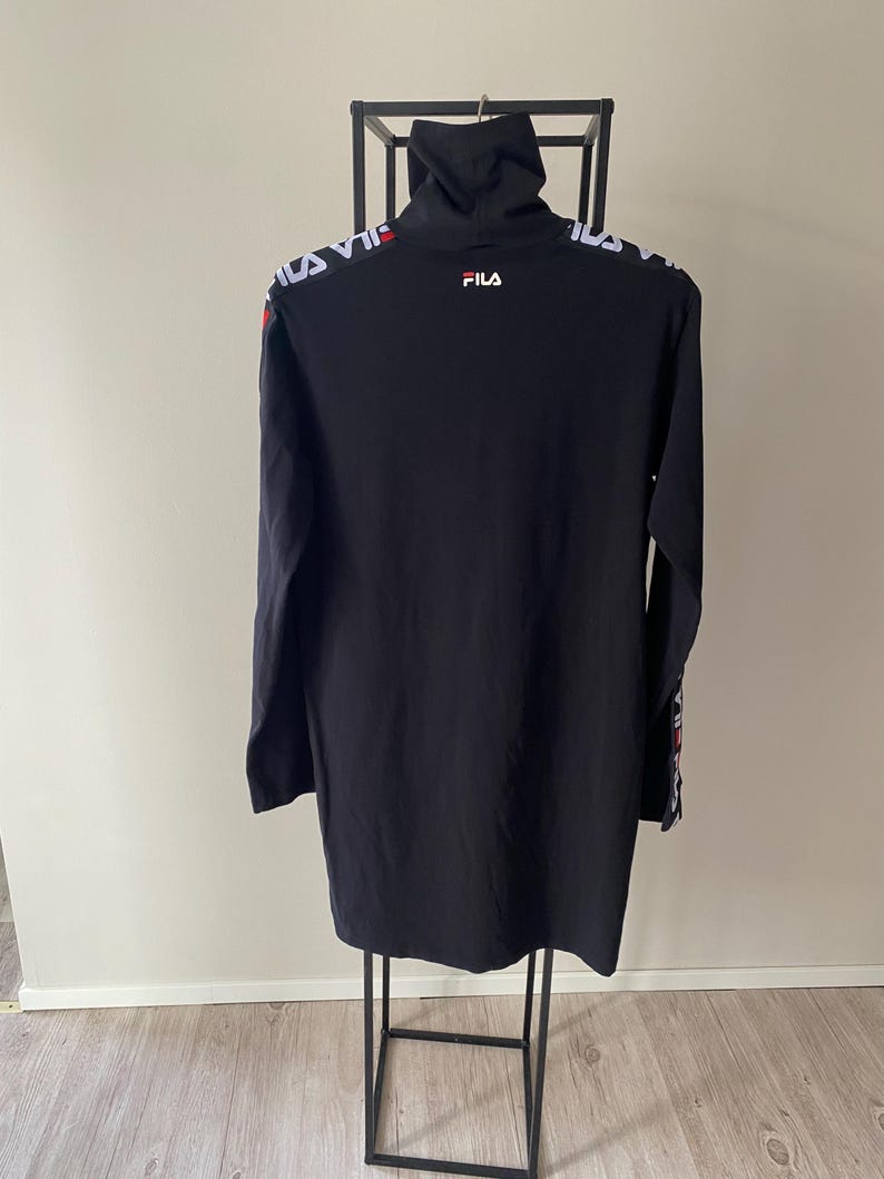 Puede incluir: Un vestido negro de manga larga con cuello alto. La prenda presenta el texto de la marca FILA en blanco y rojo en la parte superior de la espalda y a lo largo de las mangas. La tela es de color negro s&oacute;lido y el dise&ntilde;o incluye un peque&ntilde;o logotipo FILA blanco impreso en la parte superior de la espalda, debajo del cuello.