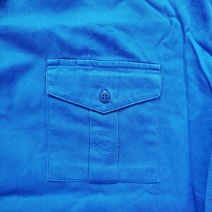 Pode incluir: Uma camisa de trabalho azul brilhante com um &uacute;nico bolso no peito e quatro bot&otilde;es. O bolso tem uma aba e um fecho de bot&atilde;o.