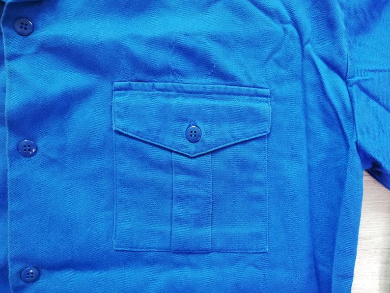 Pode incluir: Uma camisa de trabalho azul brilhante com um &uacute;nico bolso no peito e quatro bot&otilde;es. O bolso tem uma aba e um fecho de bot&atilde;o.