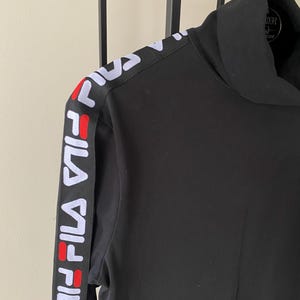 Puede incluir: Una camiseta negra de manga larga con cuello alto. Presenta una franja vertical negra a lo largo de la manga con el texto FILA en blanco repetido, acentuado con un peque&ntilde;o detalle rectangular rojo entre las letras. Tiene cuello alto.