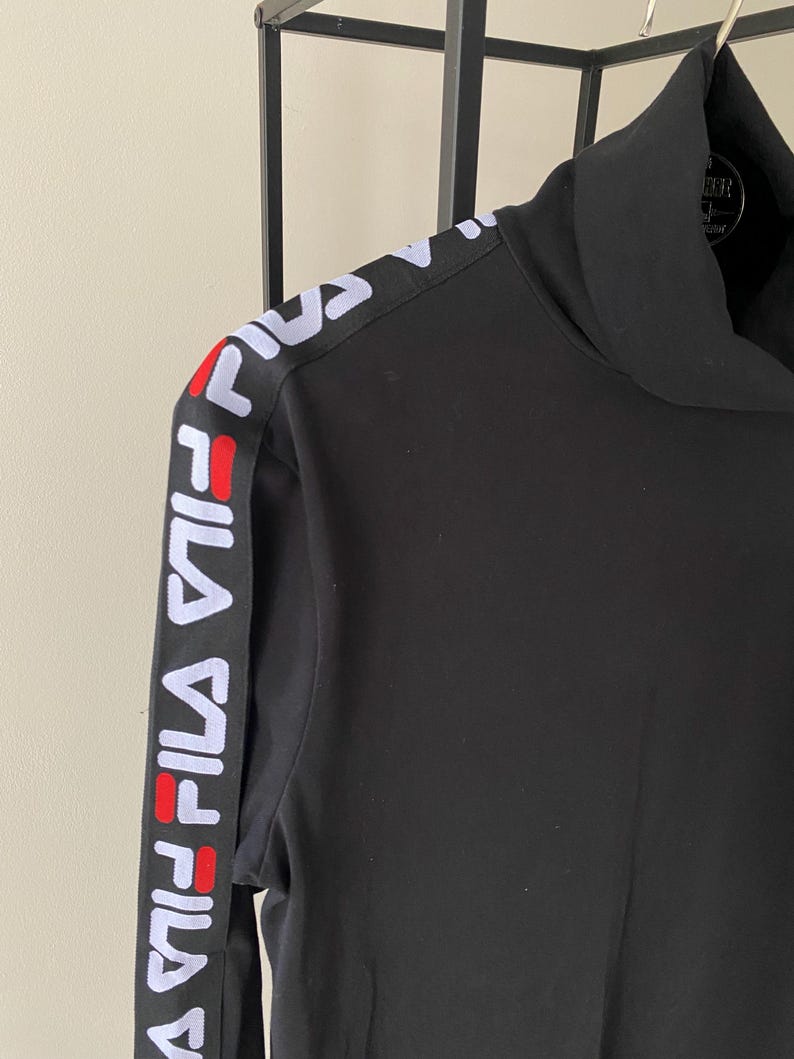 Puede incluir: Una camiseta negra de manga larga con cuello alto. Presenta una franja vertical negra a lo largo de la manga con el texto FILA en blanco repetido, acentuado con un peque&ntilde;o detalle rectangular rojo entre las letras. Tiene cuello alto.