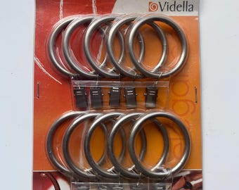 Peut inclure: Un paquet de dix anneaux de rideaux en métal argenté avec pinces, disposés en deux rangées sur un support en carton orange et blanc. L'emballage porte le logo Vidella et l'inscription 28, indiquant un diamètre de 28 mm.