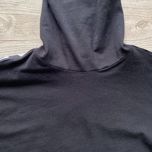 Puede incluir: Una camiseta negra de cuello alto confeccionada en tela negra s&oacute;lida con un dise&ntilde;o de cuello alto doblado. La prenda presenta una est&eacute;tica limpia con mangas largas y un detalle de raya blanca visible a lo largo de la costura del hombro.