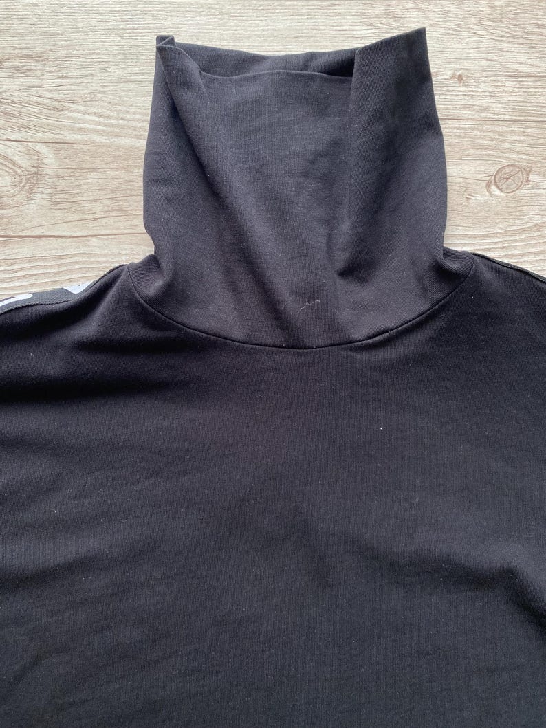 Puede incluir: Una camiseta negra de cuello alto confeccionada en tela negra s&oacute;lida con un dise&ntilde;o de cuello alto doblado. La prenda presenta una est&eacute;tica limpia con mangas largas y un detalle de raya blanca visible a lo largo de la costura del hombro.