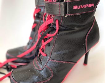Peut inclure: Une paire de bottines noires à talon aiguille, dotées de surpiqûres rose vif et de lacets roses assortis. Les bottines possèdent une bride auto-agrippante à la cheville avec le mot BUMPER brodé en fil rose sur le côté extérieur.