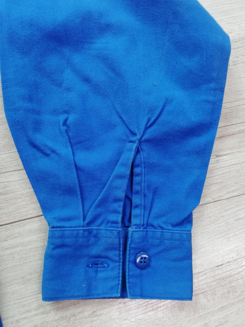Pode incluir: Close-up de uma camisa azul de manga comprida com punho de bot&atilde;o. O punho tem um &uacute;nico bot&atilde;o e uma pequena fenda na lateral.