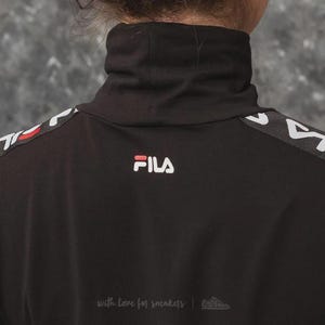 Puede incluir: Una camiseta negra de manga larga con cuello alto y el logotipo de FILA en blanco y rojo centrado bajo el cuello. Los hombros y las mangas presentan una franja negra con el nombre de la marca FILA en blanco repetido y l&iacute;neas rojas.