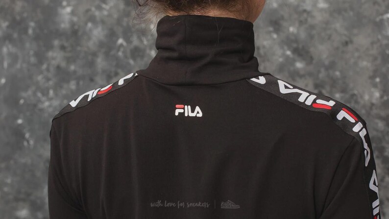 Puede incluir: Una camiseta negra de manga larga con cuello alto y el logotipo de FILA en blanco y rojo centrado bajo el cuello. Los hombros y las mangas presentan una franja negra con el nombre de la marca FILA en blanco repetido y l&iacute;neas rojas.