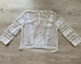 Peut inclure: Un haut à manches longues en voile blanc cassé orné de panneaux de dentelle blanche complexe disposés verticalement sur le devant et horizontalement sur les manches. Le vêtement présente une encolure ronde et une application en dentelle.
