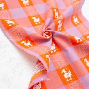 May include: A woven fabric featuring a vibrant orange and light purple gingham check pattern. The textile is decorated with small, repeating white pixelated duck motifs and subtle zigzag stitch details within the squares. The word FABLEISM is printed.