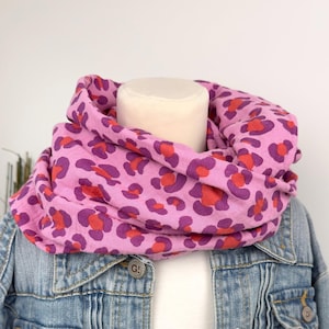 Könnte beinhalten: Ein pinkfarbener Loop-Schal mit Leopardenmuster in Lila und Rot. Der Schal wird mit einer hellblauen Jeansjacke und einem weißen Hemd getragen. Der Schal ist über einem Schaufensterpuppenkopf drapiert.