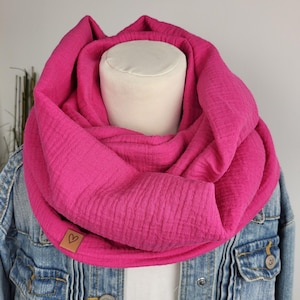 Könnte beinhalten: Ein leuchtend pinkfarbener Loop-Schal aus strukturiertem Stoff. Der Schal ist um eine Schaufensterpuppe drapiert und zeigt sein voluminöses Design. Ein kleines braunes Lederetikett mit einem Herz ist angebracht. Der Schal wird mit einer Jeansjacke kombiniert.