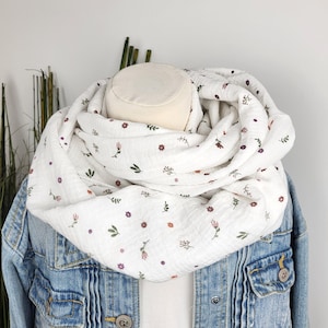 Könnte beinhalten: Ein weißer Rundschal mit Blumenmuster, drapiert über einer Jeansjacke. Der Schal zeigt kleine, sich wiederholende Designs aus lila und rosa Blumen mit grünen Blättern. Die Jacke hat eine hellblaue Waschung und sichtbare Taschen.