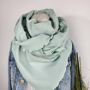 Könnte beinhalten: Ein hellgrüner Baumwollschal mit ausgefransten Kanten. Der Schal ist gefaltet und über eine Jeansjacke gelegt.
