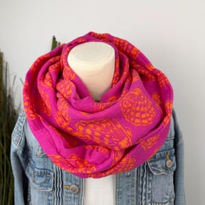 Könnte beinhalten: Ein leuchtend pinkfarbener Loop-Schal mit einem sich wiederholenden orangefarbenen Muschelmuster. Der Schal ist über einer Schaufensterpuppe drapiert und mit einer Jeansjacke kombiniert. Ein modisches Accessoire.