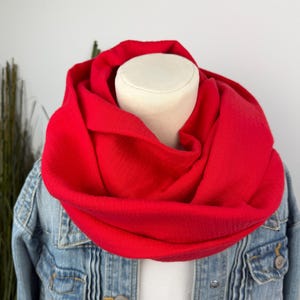 Könnte beinhalten: Ein leuchtend roter Rundschal, der um den Hals einer Schaufensterpuppe drapiert ist. Der Schal besteht aus einem weichen, strukturierten Stoff und wird mit einer Jeansjacke kombiniert. Die leuchtende Farbe des Schals setzt einen Farbakzent.