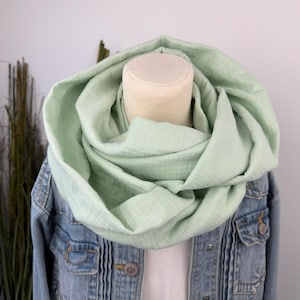Könnte beinhalten: Hellgrüner Loop-Schal, drapiert über einer Jeansjacke. Der Schal besteht aus einem weichen, strukturierten Stoff und ist um einen Schaufensterpuppenkopf gewickelt. Die Jacke hat Knopfverschlüsse und Taschen.