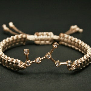 Peut inclure: Un bracelet tress&eacute; beige avec un charme en or repr&eacute;sentant une constellation de petits strass transparents.