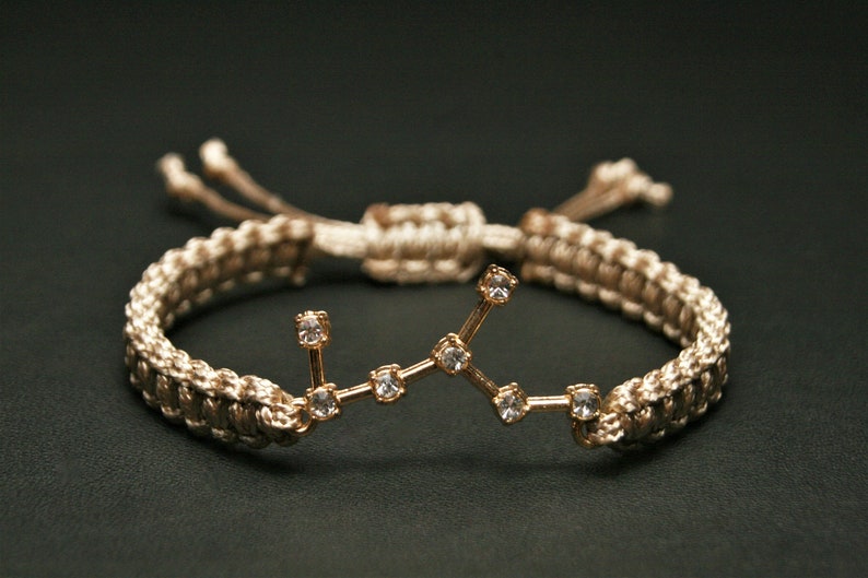 Peut inclure: Un bracelet tress&eacute; beige avec un charme en or repr&eacute;sentant une constellation de petits strass transparents.
