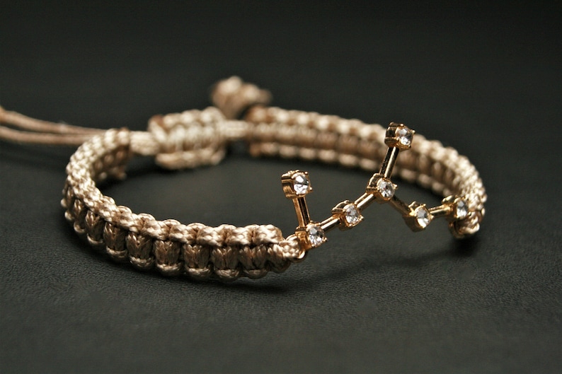 Peut inclure: Un bracelet tress&eacute; beige avec un charme de constellation dor&eacute;e orn&eacute; de strass transparents.