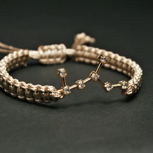 Peut inclure: Un bracelet tress&eacute; beige avec un charme de constellation dor&eacute;e orn&eacute; de petits strass transparents.