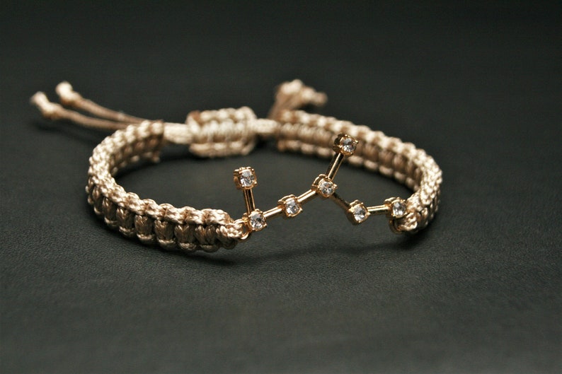 Peut inclure: Un bracelet tress&eacute; beige avec un charme de constellation dor&eacute;e orn&eacute; de petits strass transparents.