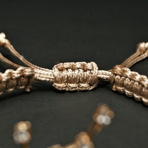 Peut inclure: Un bracelet tress&eacute; beige avec une fermeture nou&eacute;e. Le bracelet est fait d'un cordon &eacute;pais et tiss&eacute; et a un design simple et &eacute;l&eacute;gant.