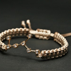 Peut inclure: Un bracelet tress&eacute; beige avec un charme en or repr&eacute;sentant la constellation de la Vierge avec des strass clairs.