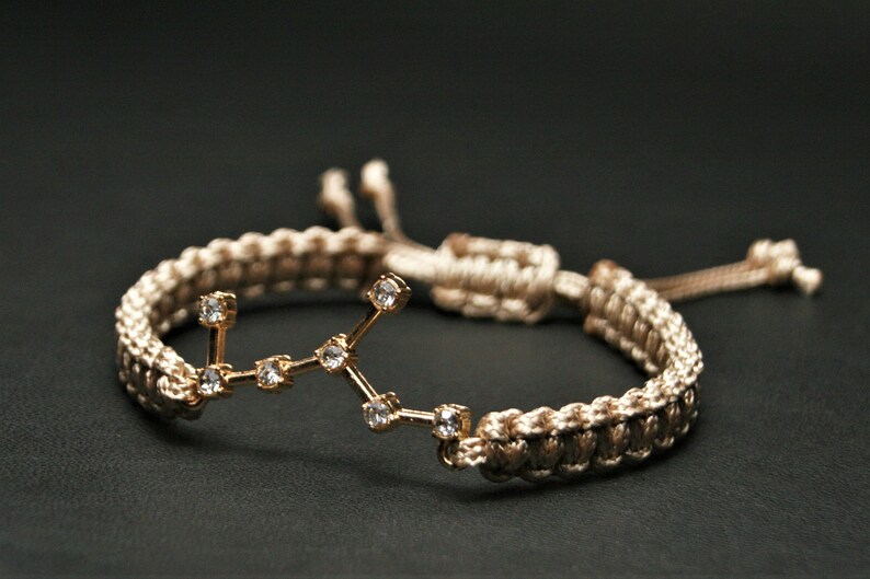Peut inclure: Un bracelet tress&eacute; beige avec un charme en or repr&eacute;sentant la constellation de la Vierge avec des strass clairs.