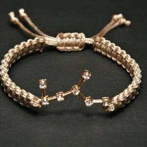 Peut inclure: Un bracelet tress&eacute; beige avec un charme de constellation Vierge en or avec des strass clairs.